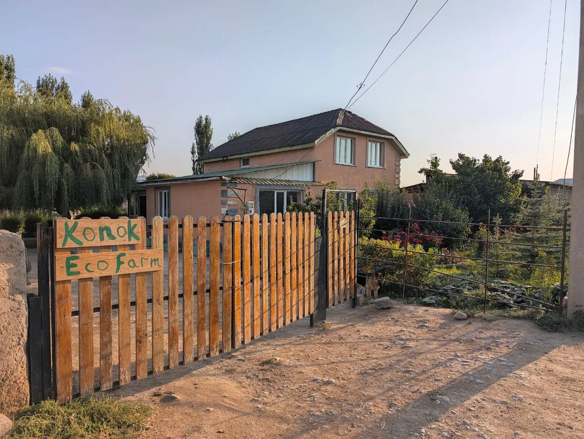 The Konok Eco Farm sign on the wooden gate, with our house and rose garden behind