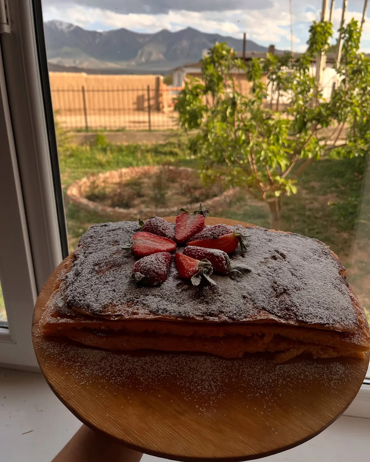 A layered cake dusted with sugar and topped with strawberries, by a window looking out to the mountains